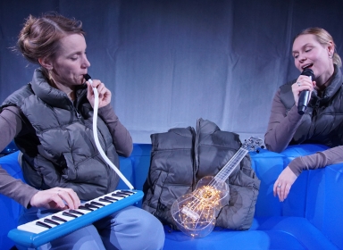 Forestillingen Usynlig, 2 kvinder ved sofa, en med melodika og 1 med håndholdt mikrofon.